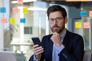 Modern ofis ortamında akıllı telefon kullanan olgun bir iş adamı. Düşünceli, gözlüklü ve koyu renk takım elbiseli, renkli yapışkan notlarla çevrili..