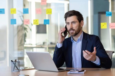 Modern ofis ortamında konuşurken cep telefonu kullanan profesyonel bir işadamı. Laptop ve yapışkan notlar görünür.