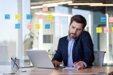 Sakallı, parlak bir ofis masasında akıllı telefon kullanan, camdaki renkli yapışkan notlarla çevrili bir iş adamı..