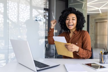 Neşeli bir Afro-Amerikalı iş kadını, bir zarf tutarken, masasında otururken, bir yumrukla kutlama yapıyor..