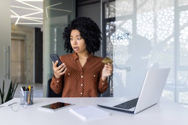 Ofis masasında şok olmuş genç bir siyahi kadın elinde kredi kartıyla akıllı telefonuna bakıyor muhtemelen beklenmedik bir işlem ile uğraşıyor..