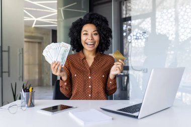 Neşeli Afrikalı Amerikalı iş kadını elinde para ve altın bir kredi kartı tutuyor, modern ofis masasında başarısını gösteriyor. Ekonomik bağımsızlığın ve başarının ifadesi.
