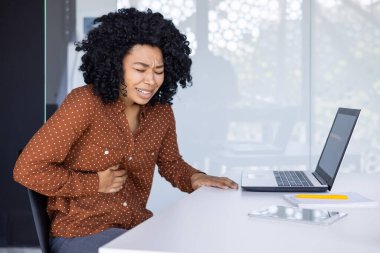 Genç bir Afrikalı Amerikalı kadın, laptopuyla modern ofis ortamında çalışırken midesi ağrıyormuş..
