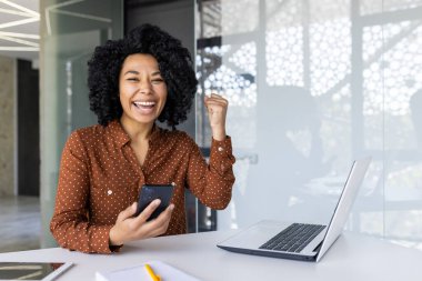 Neşeli bir Afrikalı genç Amerikalı kadın, elinde akıllı bir telefonla modern bir ofiste oturarak başarıyı kutluyor..