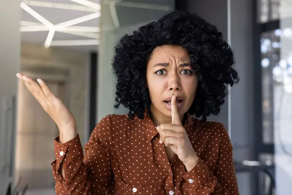 stock image A young woman with curly black hair gestures for silence in a bright, contemporary office environment. Expression of surprise and confidentiality.
