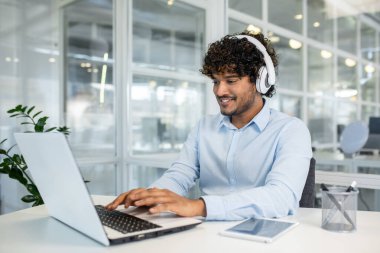 Kıvırcık saçlı neşeli bir genç, modern ofis ortamında dizüstü bilgisayar kullanarak işe odaklanırken kulaklık takıyor ve verimlilik ve konsantrasyon gösteriyor..