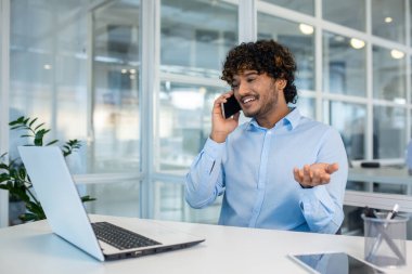 Portatif bilgisayar ekranına bakarken cep telefonuyla konuşan mutlu ofis yöneticisi. Açık mavi tişörtlü rahat bir erkek. Çalışma saatleri boyunca hafif ofiste kişisel mobil cihazlar kullanıyor..