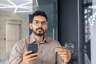 Ciddi sakallı bir adam elinde cep telefonu ve kredi kartı taşırken kameraya bakıyor. Odaklanmış banka çalışanı, İnternet 'te veri güvenliğinin korunmasının önemi üzerinde yoğunlaşıyor.