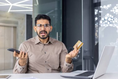 Gözlüklü kafası karışmış bir erkek cep telefonu ve plastik kartla kameraya bakarken elini yayıyor. Çalışma alanında modern ödeme seçeneklerini kullanmakla uğraşan cahil tüketici.