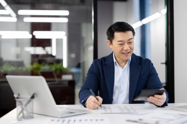 Gülümseyen Çinli muhasebeci masrafları ve geliri hesap makinesi kullanarak ve göstergeleri yazarak özetliyor. Ofiste yoğun bir gün boyunca belgeyi masalarla dolduran özenli ekonomist.