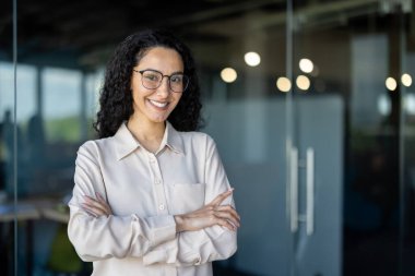 Mutlu ve başarılı iş kadını portresi, tişörtlü patron kollarını kavuşturup ofisin içinde kameraya bakıyor, kıvırcık saçlı İspanyol kadın..