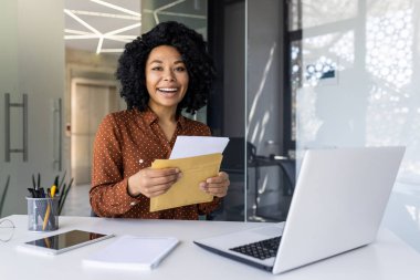 Afro-Amerikalı iş kadını, modern ofis ortamında bir zarfı açarken ışıl ışıl gülümsüyor, pozitiflik ve beklenti taşıyor..