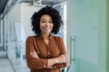 Kendine güvenen genç bir Afrikalı Amerikalı iş kadınının portresi, kollarını kavuşturmuş, parlak, modern bir ofis ortamında gülümsüyor. Şirket ve çeşitlilik temaları için mükemmel.