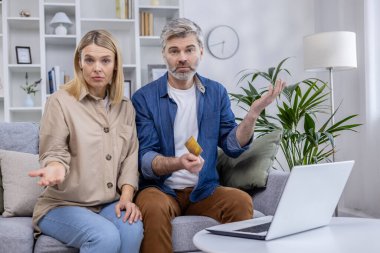 Orta yaşlı çift oturma odalarında kredi kartı sorunları ve finans konularını tartışırken endişeli görünüyor, şaşkınlık ve endişe ifadeleri sergiliyor.