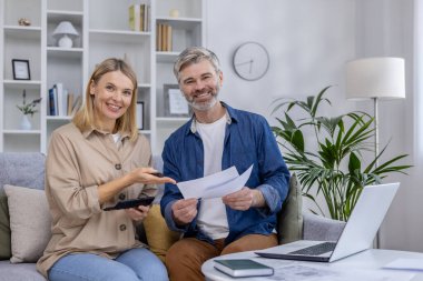 Olgun bir çift koltukta oturmuş, belgelerini ve finansal planlarını tartışırken gülümsüyor, hesap makinesi ve laptop ile modern bir oturma odasında..