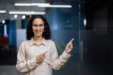 Profesyonel bir iş kadını, kıvırcık saçlı, gülümseyen ve yanında yer alan modern ofis ortamında kendine güveni ve pozitifliği olan bir iş kadını..