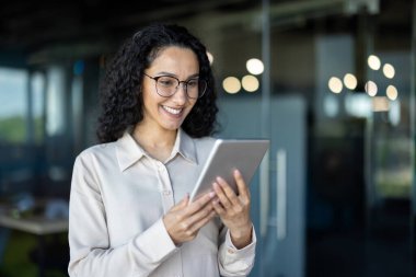 Parlak, modern ofis ortamında tablet kullanırken kıvırcık saçlı neşeli profesyonel bir kadın gülümsüyor. Görüntü bağlılık ve verimliliği iletir.