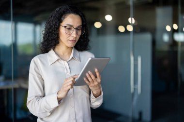 Modern ofis ortamında dijital tablet kullanan İspanyol işkadını ciddi ve düşünceli görünüyor..