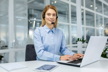 Açık mavi gömlek giyen ve kulaklık takan profesyonel bir kadın beyaz bir ofis masasında oturuyor ve parlak, modern bir ofis ortamında dizüstü bilgisayarda yazı yazıyor..