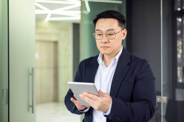 Asyalı bir işadamı tabletteki içeriği okumaya odaklanmış. Gösterişli, çağdaş bir ofis koridorunda duruyor, profesyonel bir takım elbise giymiş..