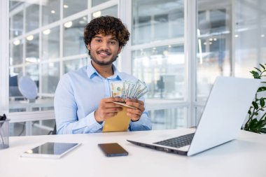 Modern bir ofiste, içi para dolu bir zarf tutarken gülümseyen genç bir iş adamı. Bu resim başarı, başarı ve finansal ödülü resmediyor..