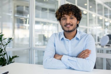 Kıvırcık saçlı, büyüleyici bir gülüşü olan parlak bir ofis ortamında oturan, kollarını kavuşturup açık mavi bir gömlek giyen genç bir adam..