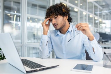 Genç yetişkin bir erkek, yanında bir akıllı telefonla, parlak ve modern bir ofiste dizüstü bilgisayarı üzerinde çalışırken stresli ve yorgun görünür..