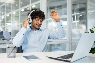 Kıvırcık saçlı, kulaklık takan neşeli bir genç adam ofis masasında dans ederken mutlu bir şekilde müzik dinliyor..