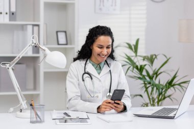 Profesyonel üniformalı siyah saçlı hemşire masasında profesyonel ekipmanlarla otururken cep telefonu ekranına bakıyor. Arap kadın davet edilen hastayı beklerken internetten mesaj atıyor.