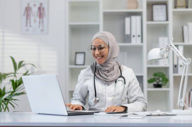 Tesettürlü neşeli bayan doktor, iyi aydınlatılmış bir ofiste masasında oturuyor ve arka planda tıbbi raporlarla dizüstü bilgisayarı üzerinde çalışıyor..