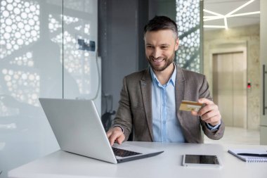 Modern ofis alanındaki neşeli iş adamı kredi kartı ve dizüstü bilgisayarla online alışveriş yapıyor. Profesyonel ve kolay e-ticaret işlemi.