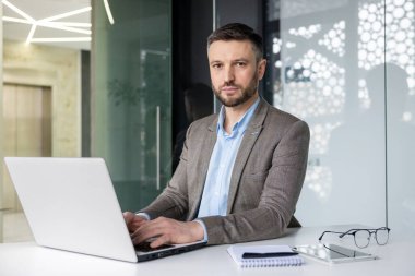 Modern ofis ortamında dizüstü bilgisayar kullanan odaklanmış bir iş adamı. Ciddi ifadesi derin bir konsantrasyon ve profesyonellik gösteriyor..