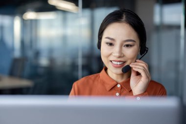 Çağdaş ofis ortamında çalışan ve gülümseyen kulaklıklı neşeli Asyalı iş kadını müşteri memnuniyetini elde ediyor..