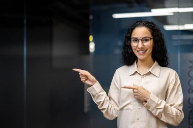 Bej gömlekli İspanyol bir iş kadını modern ofis ortamında gülümseyip onu işaret ediyor, kendine güveni ve profesyonelliği yayıyor..