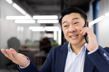 Neşeli Asyalı işadamı modern ofis ortamında bir telefon görüşmesi yaptı. Mutlu ve hevesli görünüyor, eliyle işaret ediyor..