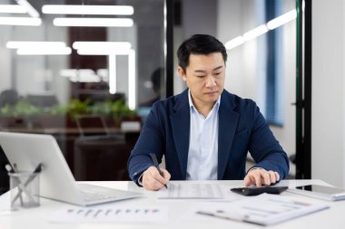 Mavi takım elbiseli profesyonel bir işadamı, grafikler ve dizüstü bilgisayarla çevrili modern bir çalışma alanındaki mali belgeleri incelemeye odaklanır..