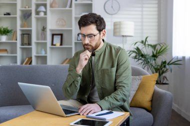 Düşünceli, orta yaşlı bir adam dizüstü bilgisayarıyla iyi dekore edilmiş bir oturma odasından çalışıyor. İfadesi, profesyonel bağlılığın ya da sorun çözmenin bir göstergesidir..