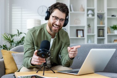 Canlı yayın yapan, kulaklıklı neşeli erkek podcaster. Konforlu evinde sıcak bir şekilde el kol hareketi yapıyor ve gülümsüyor..