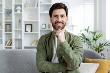 Neşeli sakallı bir adam rahat rahat oturur iyi ışıklandırılmış bir odada, bitkiler ve kitaplıklarla çevrili temiz ve modern bir iç mekan.