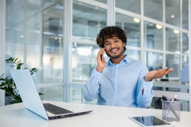 Kıvırcık saçlı, açık mavi tişörtlü neşeli genç bir adam, parlak bir ofis ortamında beyaz bir masada çalışırken akıllı bir telefonla iletişim kuruyor..