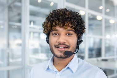 Genç bir profesyonel adam, modern ofis ortamında, müşteri desteği ve profesyonel yardımı tasvir eden bir kulaklık takarken gülümsüyor..