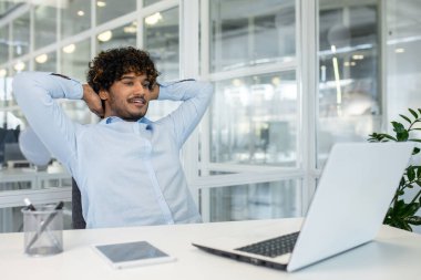 Genç bir işadamı ellerini başının arkasına koyup masasında dinleniyor, düşünceli görünüyor. Parlak ışıklandırmalı modern ofis, yoğun bir iş sırasında sakin ve düşünceli bir anı özetliyor.