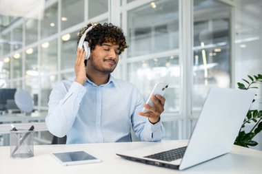 Kıvırcık saçlı genç bir adam parlak bir ofis ortamında akıllı bir telefonu tutarken kulaklıkla müzik dinliyor. İşinden memnun ve rahat görünüyor..