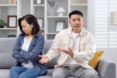 Oturma odasındaki kanepede oturan Asyalı çift. Kadın üzgün ve içine kapanık görünürken, erkek sanki bir şey açıklamaya çalışıyormuş gibi hareket ediyor, ilişkilerde anlaşmazlıklar var..