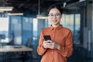 Kendine güvenen turuncu bluzlu Asyalı iş kadını modern ofis ortamında akıllı telefon kullanıyor. Profesyonel ve teknolojik temalar için mükemmel.