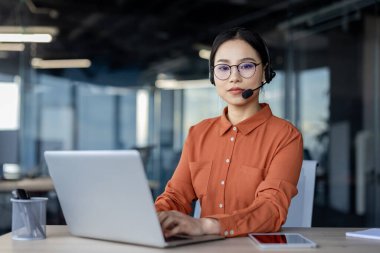 Modern ofis ortamında odaklanmış genç Asyalı kadın. Kulaklık ve gözlük takıyor, dizüstü bilgisayarına bakıyor, profesyonellik ve konsantrasyon gösteriyor..