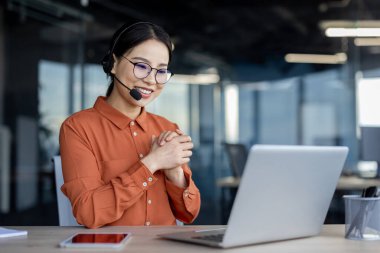 Dost canlısı Asyalı iş kadını masasında kulaklıkla modern ofis ortamında, profesyonellik ve samimiyet saçıyor..