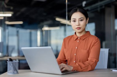 Canlı turuncu bluzlu Asyalı bir kadın ofis masasında ciddi bir şekilde oturur. Dizüstü bilgisayarı üzerinde çalışırken düşüncelere dalmış, profesyonelliği ve odaklanmayı yansıtır..