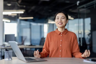 Profesyonel bir Asyalı kadın aydınlık bir ofiste huzur içinde meditasyon yapar, gözleri kapalı stresten kurtulur..