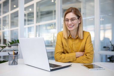 Gözlüklü genç profesyonel bir kadın dizüstü bilgisayarıyla çalışırken gülümsüyor, modern ofis ortamında..